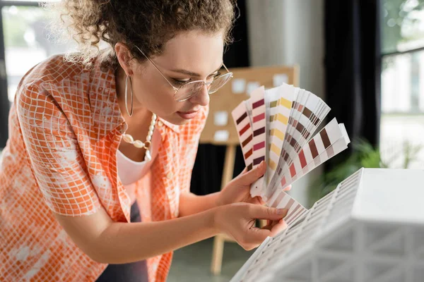 Designer de arquitetura encaracolado em óculos segurando paleta de cores perto de modelo de casa residencial no escritório — Fotografia de Stock
