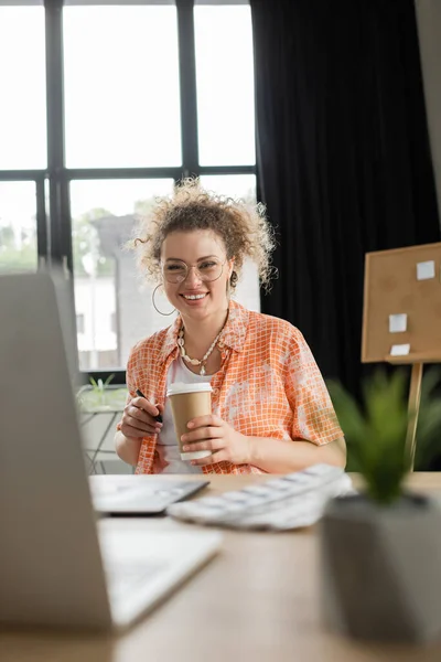 Cheerful architectural designer in glasses holding coffee to go near gadgets on desk — Stock Photo