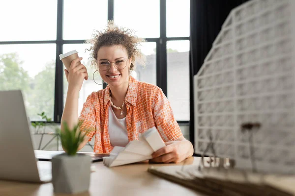 Счастливый архитектор в очках держит бумажную чашку возле ноутбука и модель жилого дома в офисе — стоковое фото