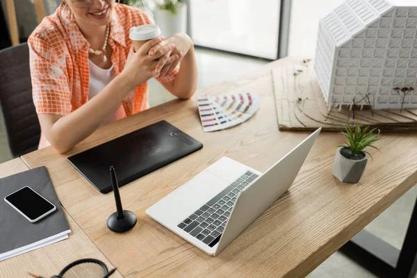 Cropped view of happy architectural designer holding coffee to go near gadgets and residential house model in office — Stock Photo