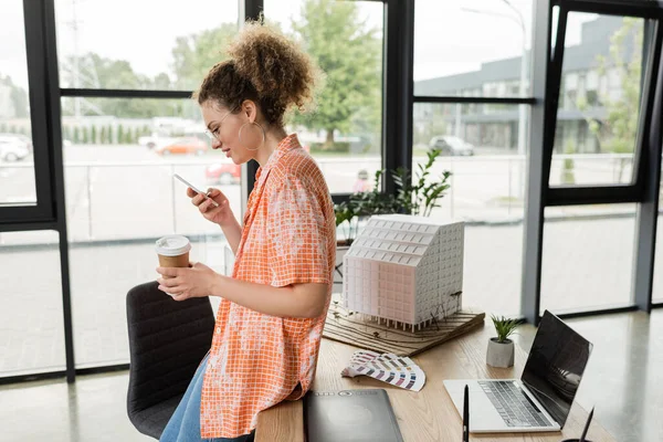Боковой вид счастливого архитектурного дизайнера в очках, держащих кофе и использующих смартфон в офисе — стоковое фото