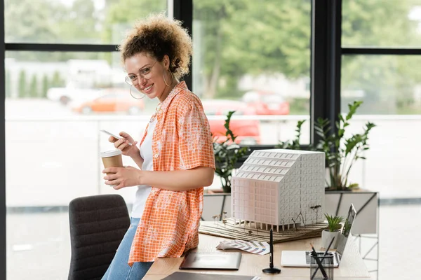 Happy architectural designer in glasses holding coffee to go and using smartphone near residential house model in office — Stock Photo