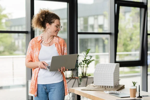 Smiling architectural designer in glasses holding laptop and looking at residential house model in office — Stock Photo