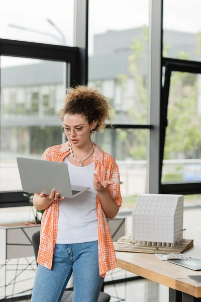 Curly architectural designer in glasses having video call on laptop near residential house model in office — Stock Photo