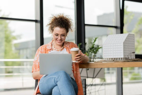 Cheerful architectural designer in glasses holding coffee to go near laptop and residential house model in office — Stock Photo