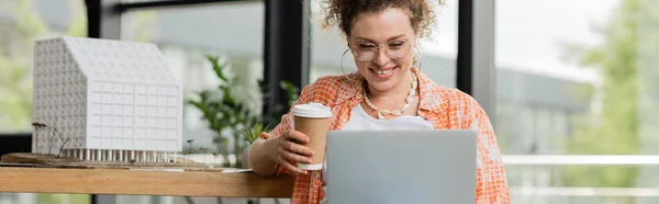 Cheerful designer in glasses holding coffee to go near laptop and residential house model in office, banner — Stock Photo