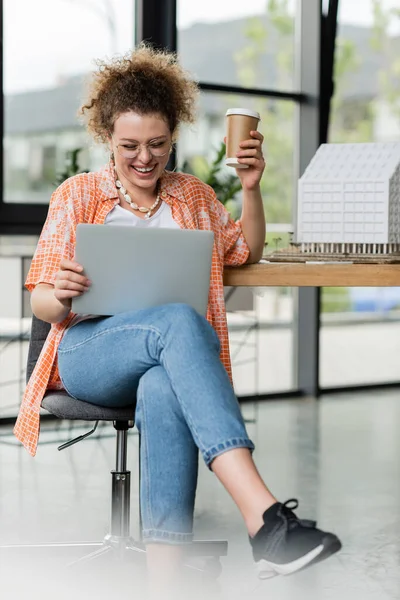 Cheerful designer in glasses holding coffee to go and looking at laptop and residential house model in office — Stock Photo