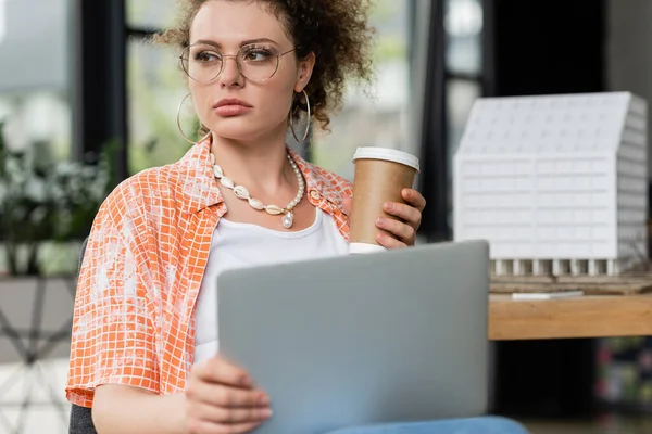 Curly designer in glasses holding coffee to go laptop near residential house model in office — Stock Photo
