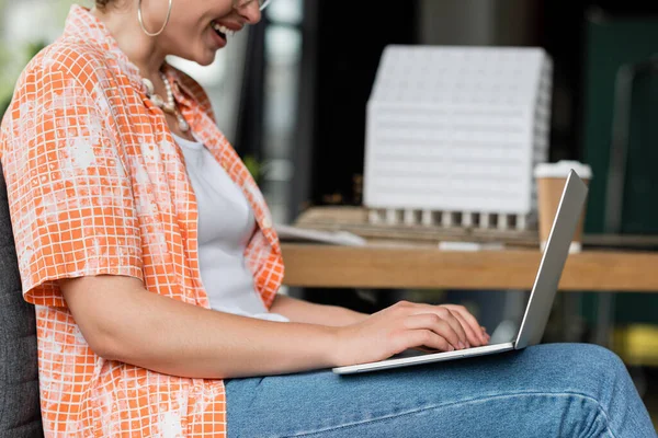 Cropped view of happy designer typing on laptop keyword near residential house model in office — Stock Photo