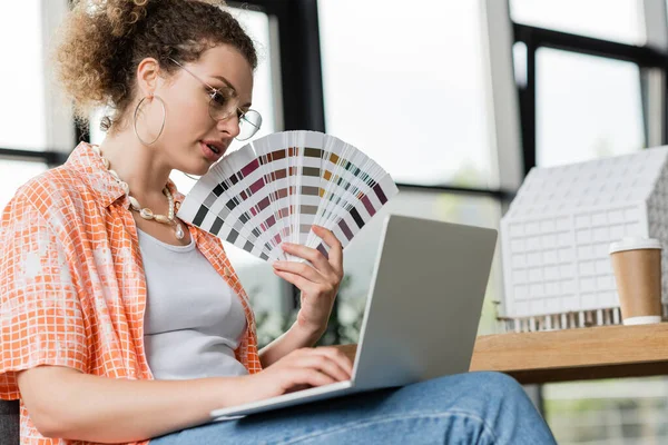 Curly architectural designer in glasses holding color palette near laptop in office — Stock Photo