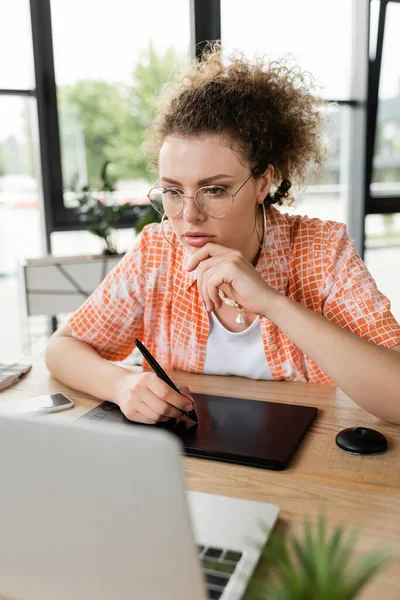 Diseñador arquitectónico rizado en gafas usando tableta gráfica y mirando el ordenador portátil mientras trabaja en la oficina - foto de stock