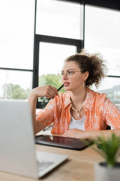 Designer arquitetônico pensivo segurando caneta estilete perto da boca enquanto sentado perto de tablet gráfico e laptop no escritório — Fotografia de Stock