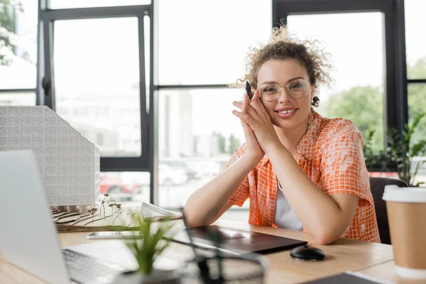 Designer sorridente in occhiali seduto vicino a gadget e modello di casa in ufficio moderno — Foto stock