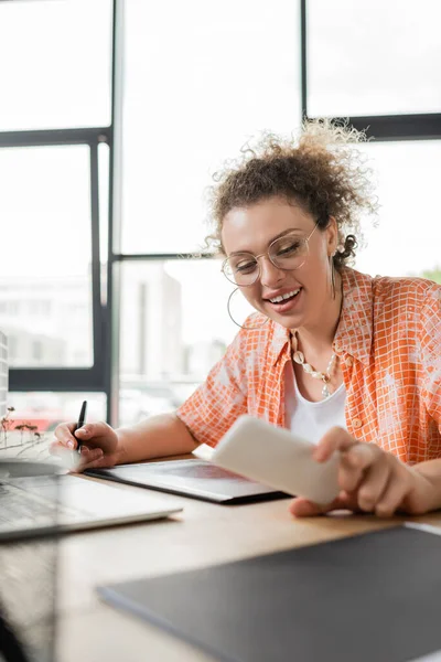 Designer feliz usando tablet gráfico e olhando para smartphone perto de laptop enquanto trabalhava no escritório — Fotografia de Stock