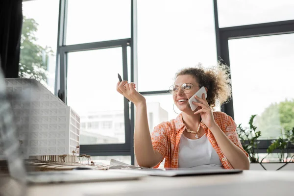Cheerful architectural designer talking on smartphone and holding stylus near residential house model — Stock Photo