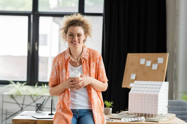 Cheerful architectural designer holding paper cup while standing near house model in office — Stock Photo