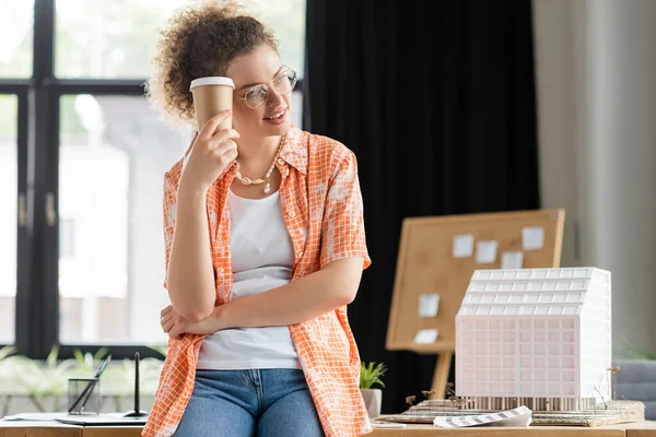 Pleased architectural designer holding paper cup while standing near house model in office — Stock Photo