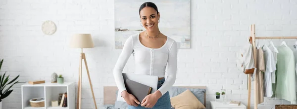 Cheerful african american student holding laptop and book in modern apartment, banner — Stock Photo