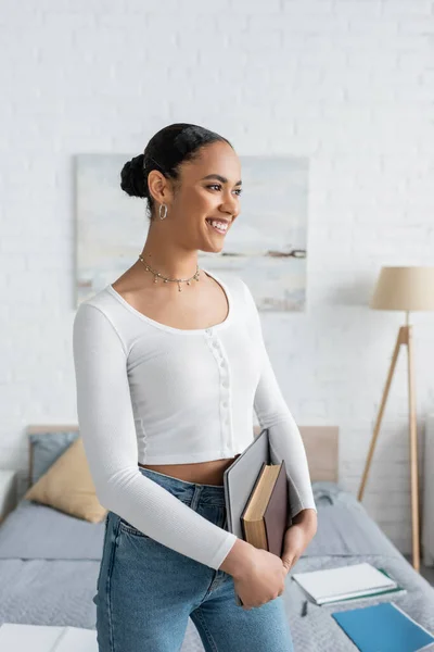Estudiante afroamericano sonriente sosteniendo portátil y reservar en apartamento moderno - foto de stock