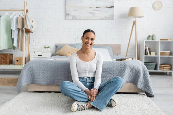 Allegro studente afroamericano in jeans seduto con le gambe incrociate vicino al letto a casa — Foto stock