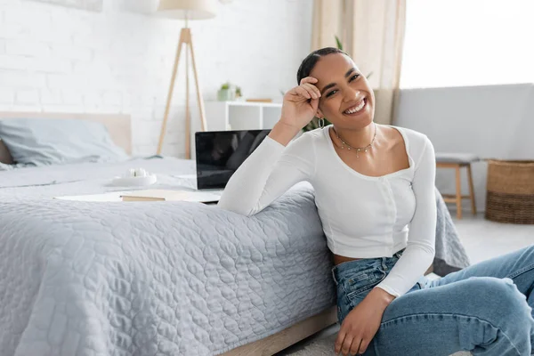 Affascinante studente afroamericano sorridente mentre seduto vicino al computer portatile sul letto — Foto stock