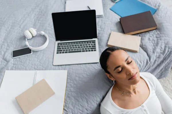 Top view of african american student with closed eyes resting near laptop and books on bed — Stock Photo