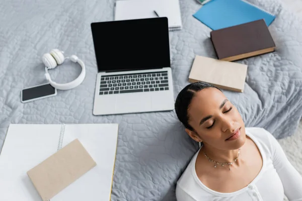 Blick von oben auf einen afrikanisch-amerikanischen Studenten mit geschlossenen Augen, der neben Laptop und Büchern auf dem Bett ruht — Stockfoto