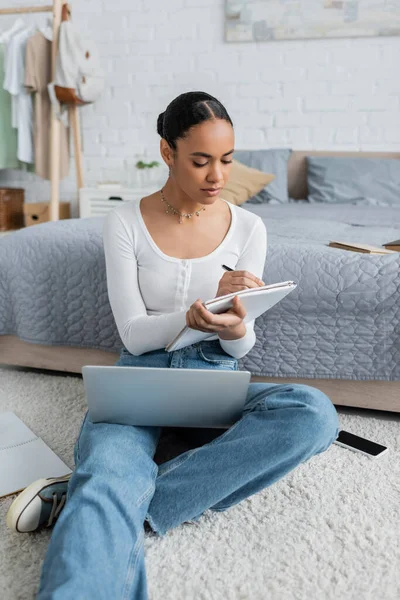 Довольно африканская американка в джинсах делает заметки во время просмотра вебинара на ноутбуке — стоковое фото