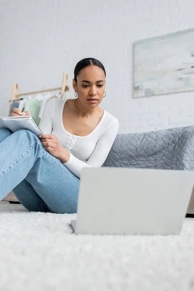 Довольно африканская американка в джинсах делает заметки во время просмотра онлайн лекции на ноутбуке — стоковое фото