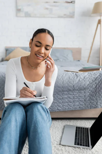 Fröhliche afrikanisch-amerikanische Frau in Jeans, die in Notizbuch schreibt, während sie auf dem Smartphone neben dem Laptop spricht — Stockfoto
