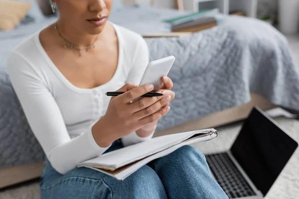 Vista recortada de la joven afroamericana utilizando un teléfono inteligente y sentado junto a la computadora portátil mientras estudia desde casa - foto de stock