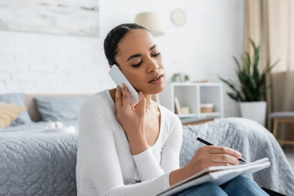 Estudiante afroamericano bastante tomar notas mientras habla en el teléfono inteligente y estudiar desde casa - foto de stock