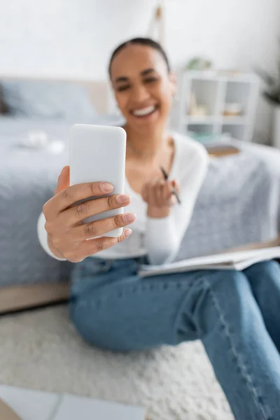 Estudiante afroamericano feliz y borrosa tomando selfie en teléfono inteligente mientras estudia desde casa - foto de stock