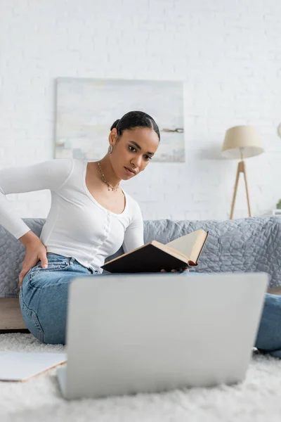 Estudiante afroamericano bastante teniendo dolor de espalda mientras sostiene el libro y mirando el ordenador portátil - foto de stock