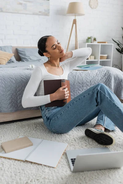 Jeune étudiant afro-américain ayant mal au cou tout en tenant un livre et en étudiant à la maison — Photo de stock