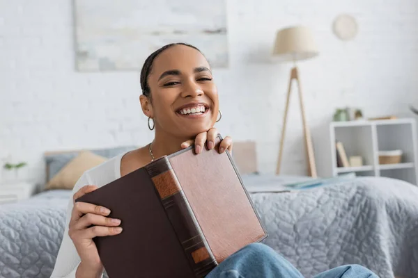 Complacido estudiante afroamericano sosteniendo libro mientras estudia desde casa - foto de stock