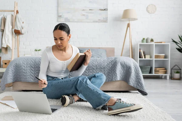 Studente afro-americano che tiene un libro mentre guarda una lezione online sul computer portatile e studia da casa — Foto stock