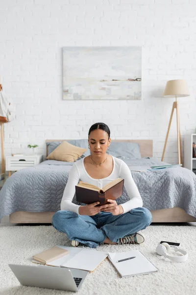 Young african american student sitting near gadgets and reading book — Stock Photo