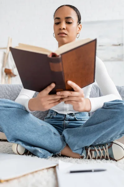 Vista de ángulo bajo del joven estudiante afroamericano sentado con las piernas cruzadas y libro de lectura - foto de stock