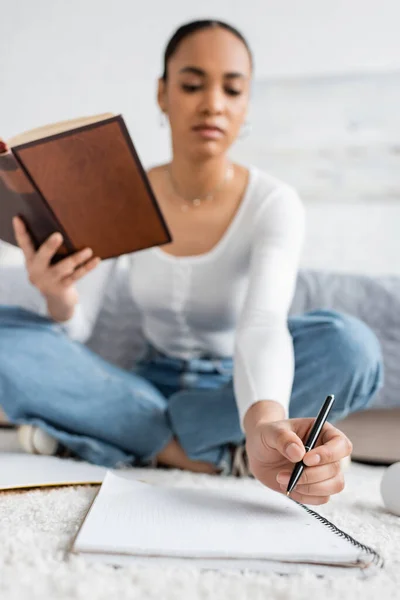 Vista a basso angolo di giovane studente afroamericano seduto con libro mentre prende appunti su notebook — Foto stock