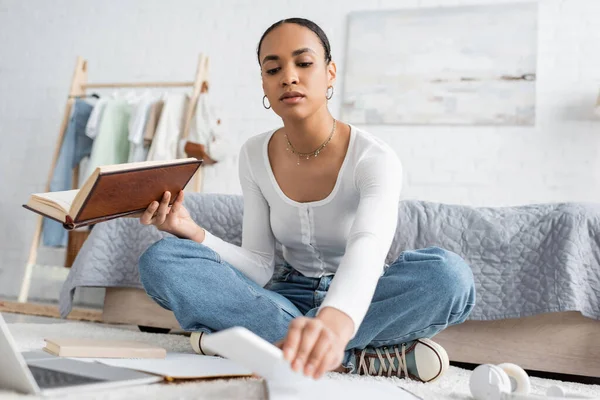 Ocupado estudiante afroamericano sosteniendo libro y tomando teléfono inteligente mientras estudia desde casa - foto de stock