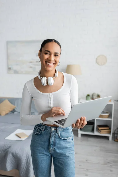 Estudante americano africano feliz com fones de ouvido sem fio no pescoço usando laptop no quarto moderno — Fotografia de Stock