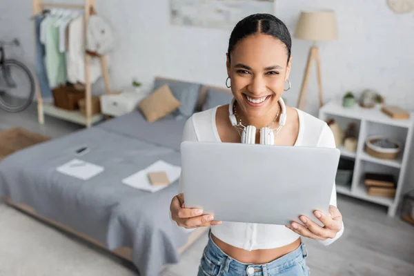 Vista aérea de estudiante afroamericano alegre con auriculares inalámbricos en el cuello usando el ordenador portátil en el dormitorio moderno - foto de stock