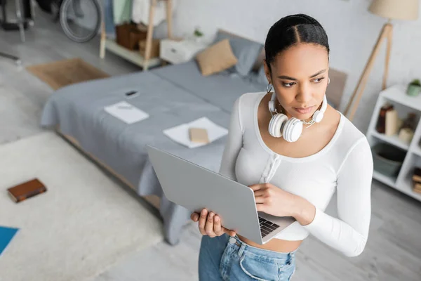 Hochwinkelaufnahme einer hübschen afrikanisch-amerikanischen Studentin mit drahtlosen Kopfhörern am Hals, die einen Laptop im modernen Schlafzimmer hält — Stockfoto