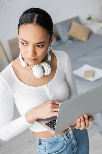 Vue grand angle de jeune étudiant afro-américain avec écouteurs sans fil sur le cou tenant ordinateur portable — Photo de stock