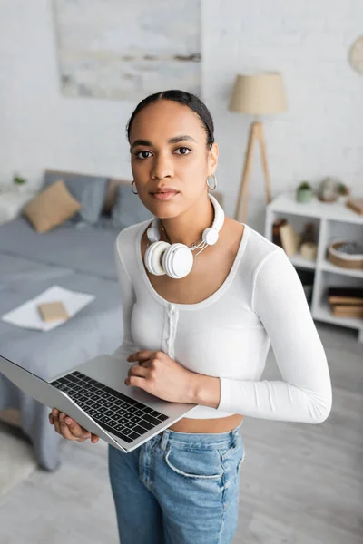 Vista aérea del estudiante afroamericano con auriculares inalámbricos en el cuello sosteniendo portátil en el dormitorio — Stock Photo