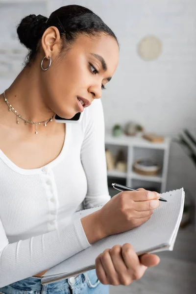 Junge afrikanisch-amerikanische Studentin spricht auf Smartphone, während sie in Notizbuch schreibt — Stockfoto