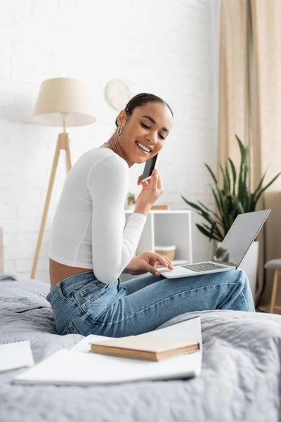 Smiling african american student talking on smartphone while using laptop and learning online from home — Stock Photo