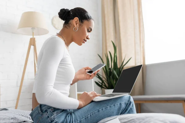 Jovem estudante afro-americano segurando smartphone e usando laptop enquanto aprende on-line de casa — Fotografia de Stock