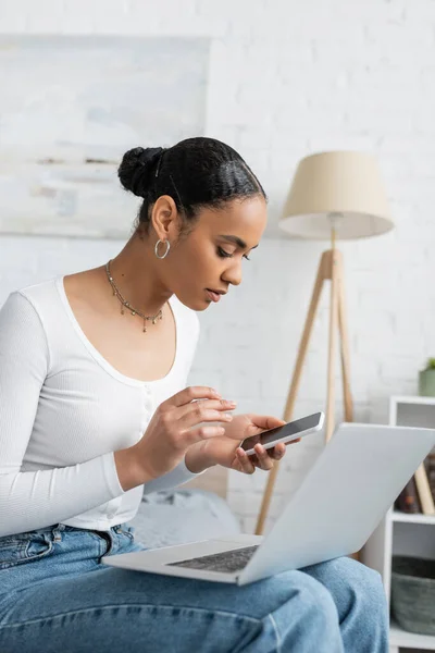 Estudiante afroamericano joven usando un teléfono inteligente cerca de la computadora portátil mientras aprende en línea desde casa - foto de stock
